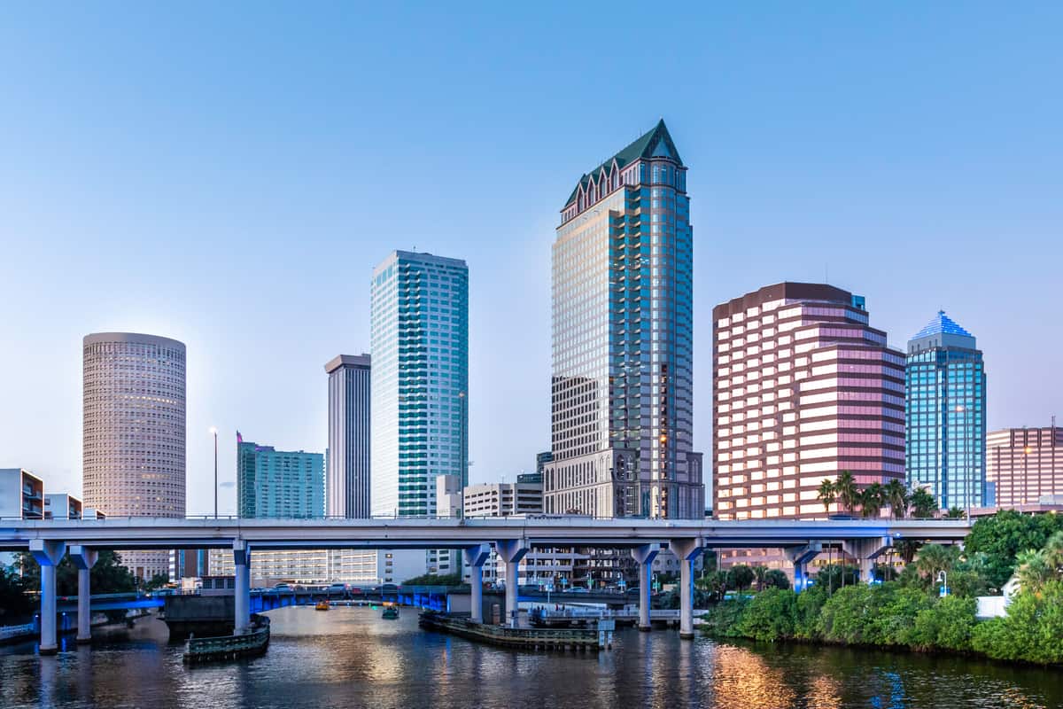 The downtown Tampa, Florida skyline at dusk seen across the Hillsborough River, with the cylindrical Rivergate Tower at left and the elevated Selmon Expressway crossing the water in the foreground